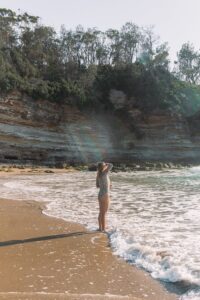 Side view of unrecognizable female in swimwear standing with raised arm on sandy beach in wavy ocean against rocky cliff in sunny day